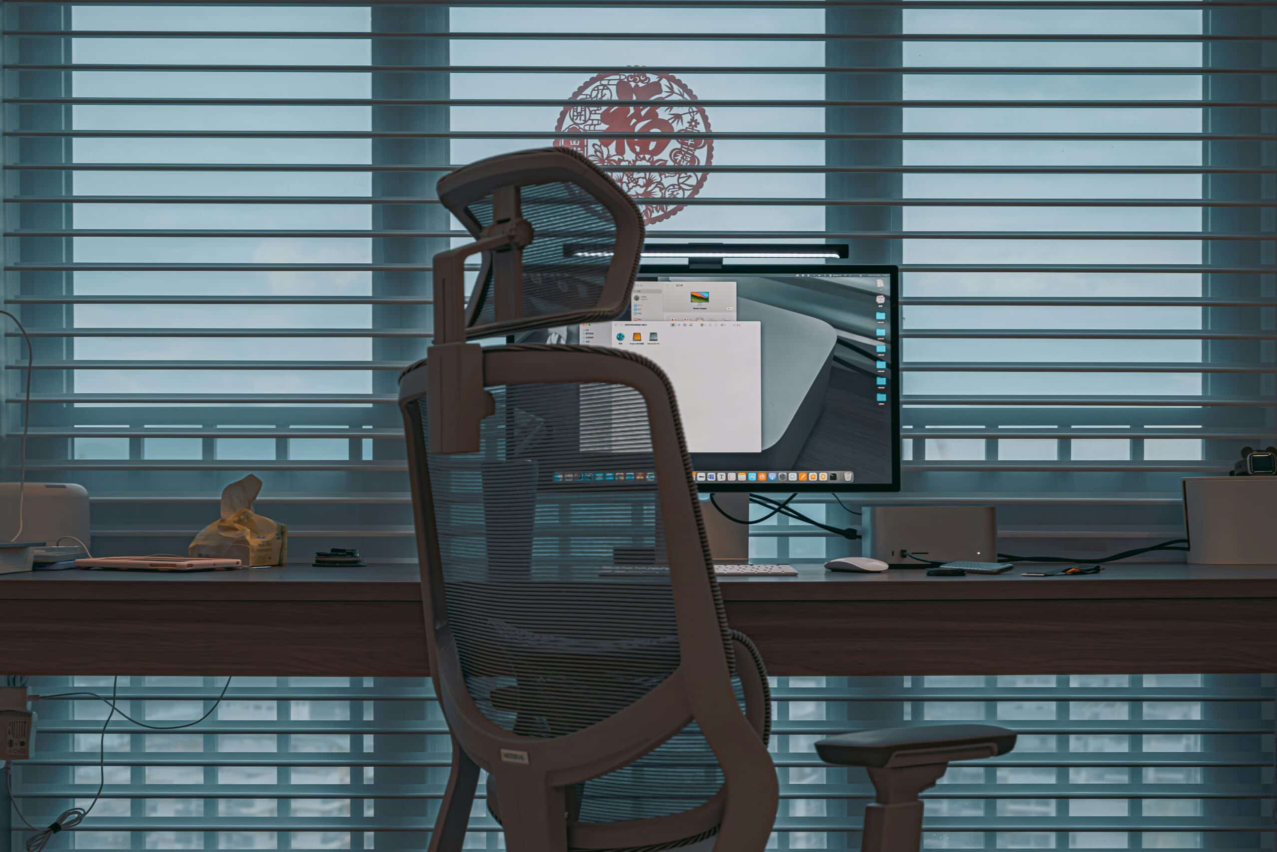 Modern office workstation with ergonomic chair and computer monitor in front of window blinds